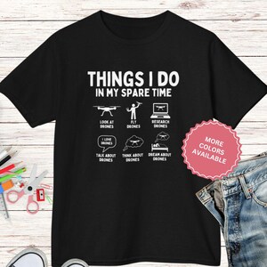 May include: Black t-shirt with white text and graphics about drones. The text reads "THINGS I DO IN MY SPARE TIME" with illustrations of drone-related activities. A pink sticker indicates "MORE COLORS AVAILABLE".