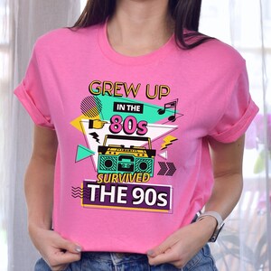 May include: Pink t-shirt with a graphic design featuring a boombox, musical notes, and the text "Grew Up in the 80s Survived the 90s".