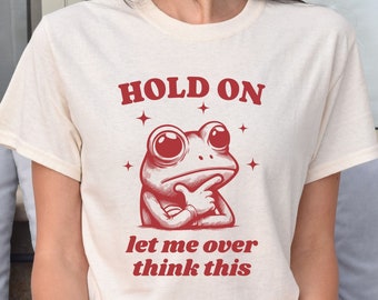 May include: A white t-shirt with a red graphic of a frog thinking. The text on the shirt reads "HOLD ON let me over think this".