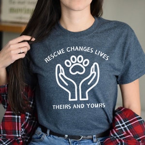May include: A dark blue t-shirt with white text that reads "Rescue Changes Lives" and a white paw print inside two hands. The text below the paw print reads "Theirs and Yours".