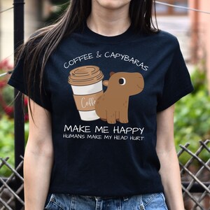 May include: Black t-shirt with a cartoon image of a capybara and a cup of coffee. The text on the shirt reads "Coffee & Capybaras Make Me Happy Humans Make My Head Hurt".