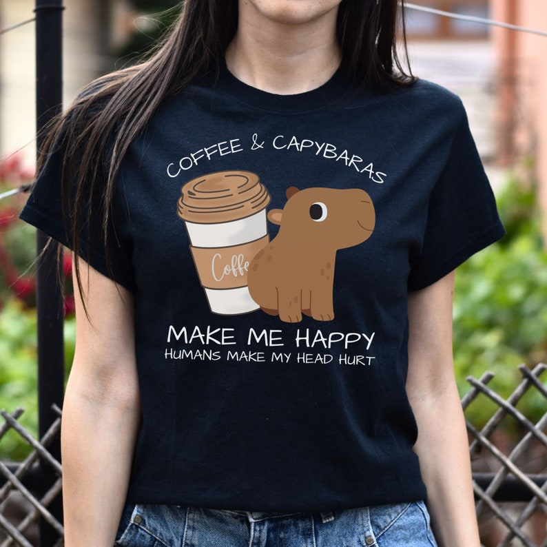 May include: Black t-shirt with a cartoon image of a capybara and a cup of coffee. The text on the shirt reads "Coffee & Capybaras Make Me Happy Humans Make My Head Hurt".
