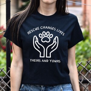 May include: Black t-shirt with a white graphic of two hands holding a paw print. The text "Rescue Changes Lives" is above the graphic and "Theirs and Yours" is below.