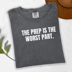 May include: A gray t-shirt with the text "THE PREP IS THE WORST PART" in white block letters. The shirt is folded and has a crew neck. A brown hat is visible in the background.