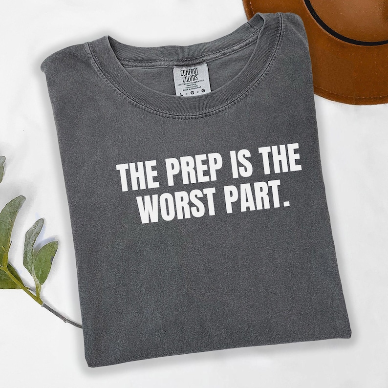 May include: A gray t-shirt with the text "THE PREP IS THE WORST PART" in white block letters. The shirt is folded and has a crew neck. A brown hat is visible in the background.