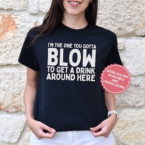 Pode incluir: T-shirt preto com texto branco: "I'M THE ONE YOU GOTTA BLOW TO GET A DRINK AROUND HERE." Um adesivo rosa à direita diz "MORE COLORS AVAILABLE AND UNISEX SIZING."