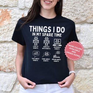 May include: A black short-sleeved t-shirt featuring white text that reads THINGS I DO IN MY SPARE TIME, accompanied by six white graphic icons of robots with labels including LOOK AT ROBOTS, HANG OUT WITH ROBOTS, RESEARCH ROBOTS, and DREAM ABOUT ROBOTS.