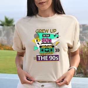 May include: A beige t-shirt with a graphic design featuring a retro boombox, the text "Grew Up in the 80s Survived the 90s", and colorful geometric shapes.