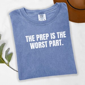 May include: A blue t-shirt with the text "THE PREP IS THE WORST PART" in white block letters. The shirt is folded and has a Comfort Colors tag. A brown hat is partially visible in the background.