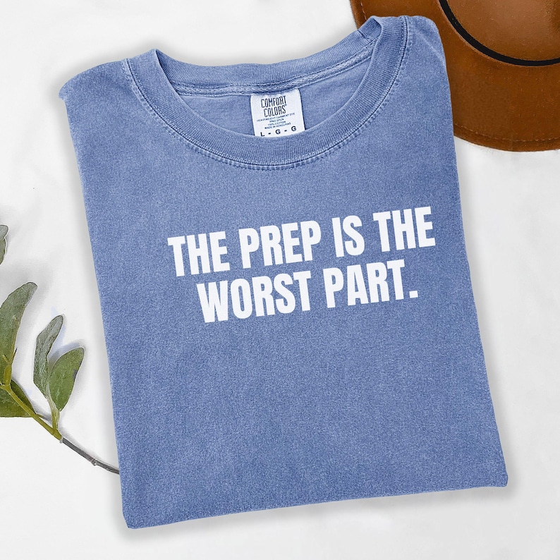 May include: A blue t-shirt with the text "THE PREP IS THE WORST PART" in white block letters. The shirt is folded and has a Comfort Colors tag. A brown hat is partially visible in the background.