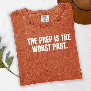 May include: Burnt orange t-shirt with the text "THE PREP IS THE WORST PART" in white block letters. The shirt is folded and has a Comfort Colors tag. A brown hat is partially visible in the background.
