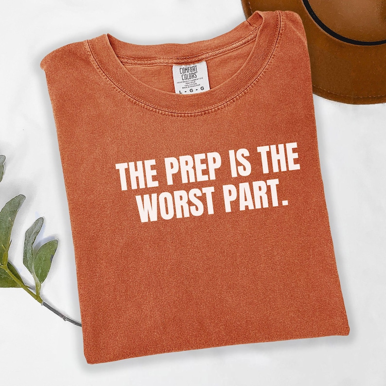 May include: Burnt orange t-shirt with the text "THE PREP IS THE WORST PART" in white block letters. The shirt is folded and has a Comfort Colors tag. A brown hat is partially visible in the background.