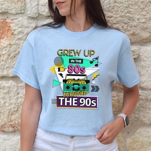 May include: Light blue t-shirt with a graphic design featuring a boombox, musical notes, and the text "Grew Up in the 80s Survived the 90s".