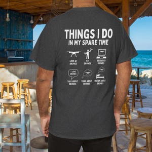 Pode incluir: Camiseta cinza escuro com texto e gr&aacute;ficos brancos sobre drones. O texto diz "THINGS I DO IN MY SPARE TIME" com ilustra&ccedil;&otilde;es de drones e atividades relacionadas como "LOOK AT DRONES" e "DREAM ABOUT DRONES."