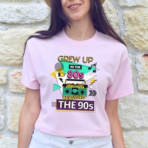 May include: Pink t-shirt with a graphic design featuring a boombox, musical notes, and the text "Grew Up in the 80s Survived the 90s".