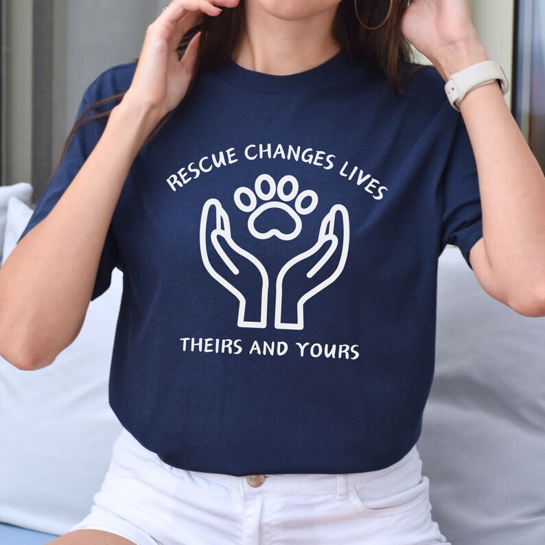 May include: Navy blue t-shirt with a white graphic of two hands holding a paw print. The text "Rescue Changes Lives" is above the graphic and "Theirs and Yours" is below.