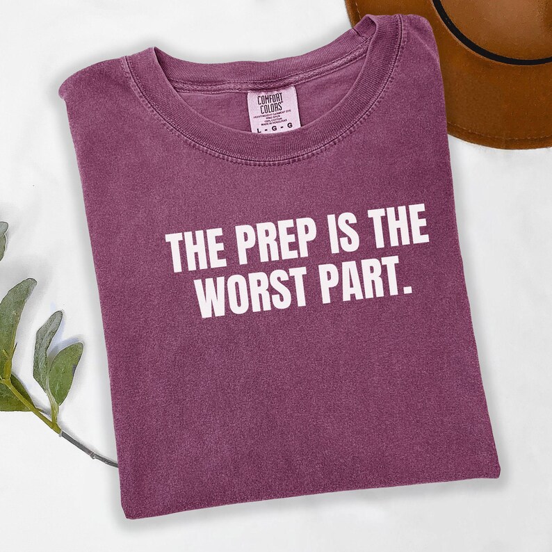 May include: A maroon Comfort Colors t-shirt with the text "THE PREP IS THE WORST PART" in white block letters. The shirt is folded and lies on a white surface. A brown hat is partially visible in the upper right corner.