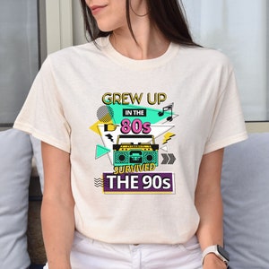 May include: A white t-shirt with a graphic design featuring a boombox, music notes, and the text "Grew Up In The 80s Survived The 90s".