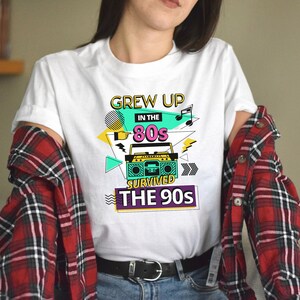 May include: White t-shirt with a graphic design featuring a boombox, the text "Grew Up in the 80s Survived the 90s", and colorful geometric shapes.