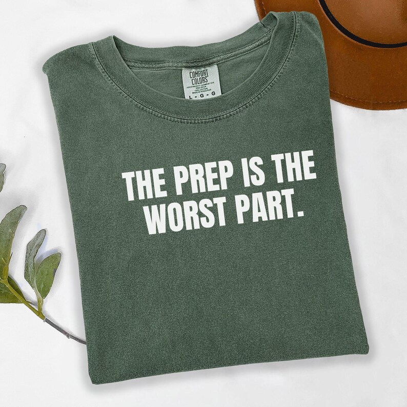 May include: Green t-shirt with the text "THE PREP IS THE WORST PART" in white. The shirt is folded and has a Comfort Colors tag. A brown hat is partially visible in the background.