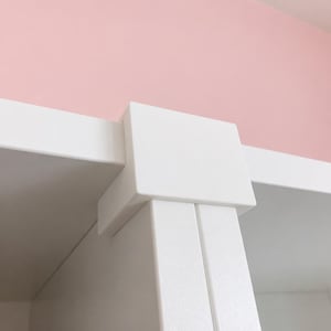 May include: Close-up of a white shelf unit corner. The unit has a clean, modern design with a square corner post and horizontal shelf supports. The background is a soft pink.