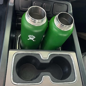 Könnte beinhalten: Zwei grüne, isolierte Wasserflaschen mit silbernen Deckeln befinden sich in einem Getränkehalter im Auto. Die Flaschen haben ein weißes Logo. Der Getränkehalter befindet sich in der Konsole des Autos.