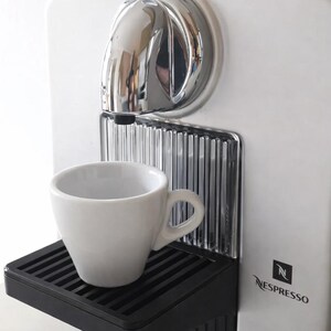May include: A white Nespresso coffee machine with a chrome spout and a black drip tray. A white ceramic coffee cup sits on the tray. The machine has the Nespresso logo on the side.