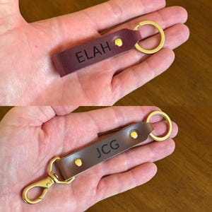 May include: Two leather keychains with gold metal rings and clasps. One keychain is brown with the name "JCG" engraved on it. The other keychain is burgundy with the name "ELAH" engraved on it.