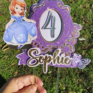 Puede incluir: Topper de pastel de purpurina morado y dorado con una princesa de dibujos animados, el número 4 y el nombre "Sophie". También se incluye una mariposa.