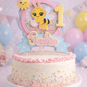 May include: A decorated cake with a bee-themed topper for a first birthday. The cake has pink frosting and colorful sprinkles. The topper features a bee, the number 1, and the name "Sophie".