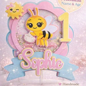 May include: A custom bee-themed birthday cake topper. The topper features a yellow bee holding a small bag, a yellow number one, and the name "Sophie" in pink. The design includes a shaker element and the text "Perfect for First Birthday".
