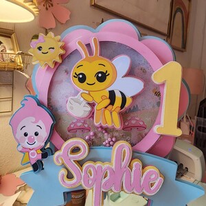May include: A layered cake topper featuring a cartoon bee holding a volleyball, a smiling sun, and a cartoon character. The topper includes the name "Sophie" and the number "1" in pink and yellow, set against a blue and pink floral design.