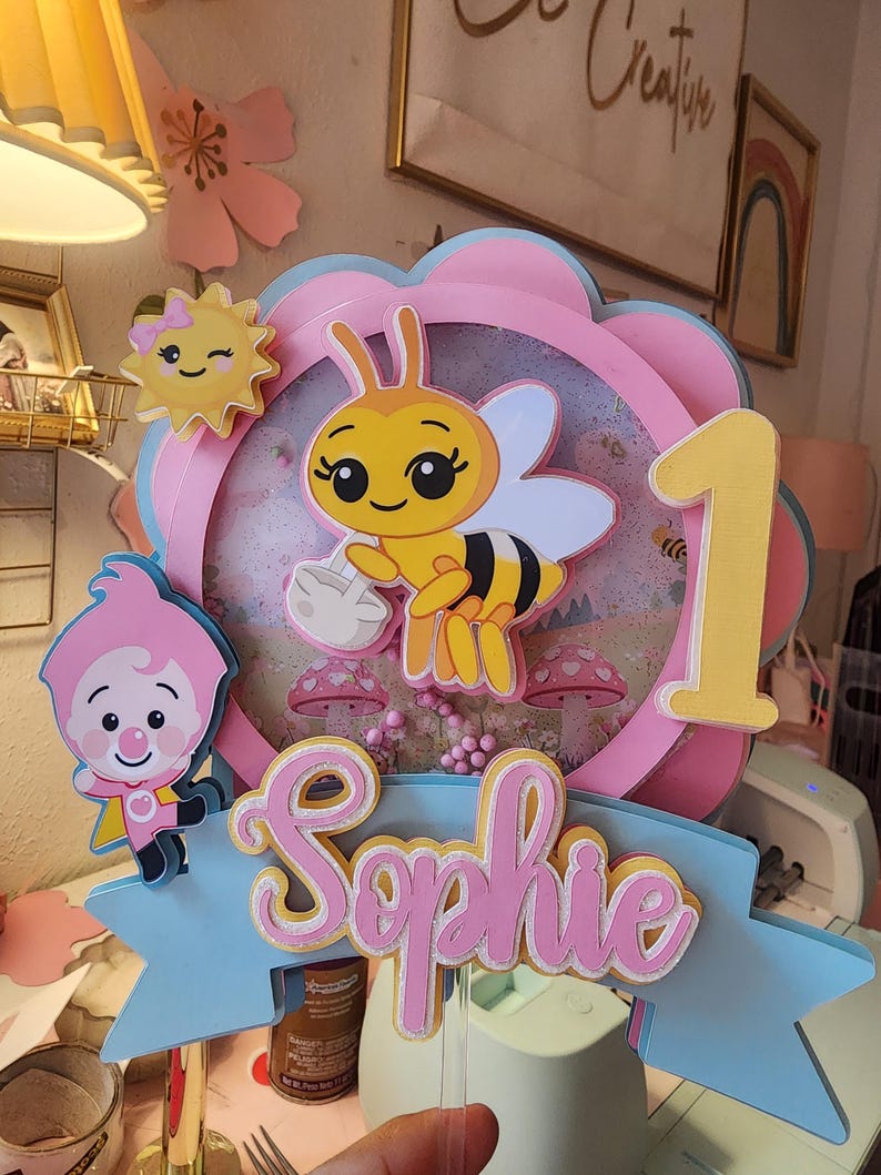 May include: A layered cake topper featuring a cartoon bee holding a volleyball, a smiling sun, and a cartoon character. The topper includes the name "Sophie" and the number "1" in pink and yellow, set against a blue and pink floral design.