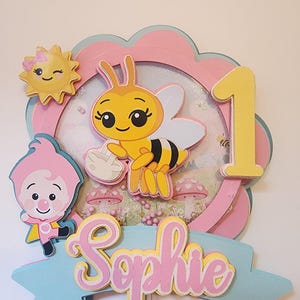 May include: A layered paper cake topper with a bee, sun, and cartoon character. The design includes the number 1, the name "Sophie", and a pink, yellow, and blue color scheme. The topper is likely for a first birthday celebration.
