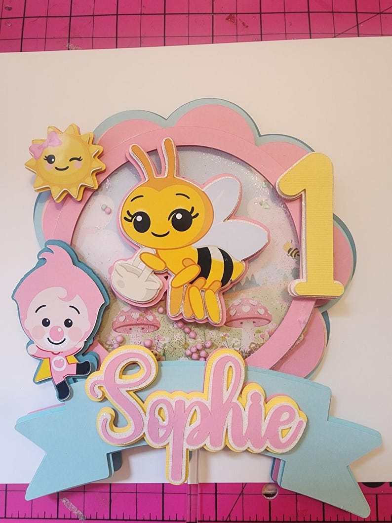 May include: A layered paper cake topper with a bee, sun, and cartoon character. The design includes the number 1, the name "Sophie", and a pink, yellow, and blue color scheme. The topper is likely for a first birthday celebration.