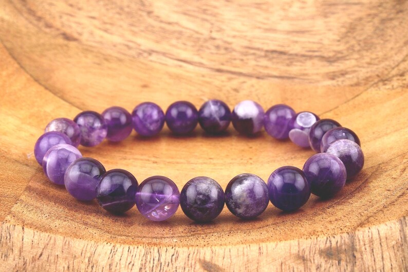 May include: A purple amethyst gemstone bracelet with round beads. The bracelet is on a wooden surface.