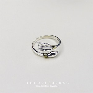 May include: A silver ring with an arrow design. The arrow has feather detailing and is accented with gold-coloured bands. The ring is set against a white background. The text "THEUSEFULBAG Handcrafted Jeweller" is at the bottom.