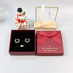 May include: An assortment of jewelry and decorative items. Includes a red gift box with a bow, earrings, a ring, a snowman figurine, and a necklace on a display stand. The gift box has the text "BEST WISHES FOR YOU Especially for you."