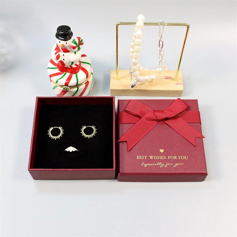 May include: An assortment of jewelry and decorative items. Includes a red gift box with a bow, earrings, a ring, a snowman figurine, and a necklace on a display stand. The gift box has the text "BEST WISHES FOR YOU Especially for you."