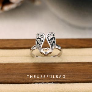 Op de afbeelding: Een zilveren ring met een engelenvleugelontwerp en een hartdetail. De ring heeft gedetailleerde veertjes en wordt gepresenteerd op een houten oppervlak. De tekst "THEUSEFULBAG" en "Handcrafted Jewellery" staan onderaan.