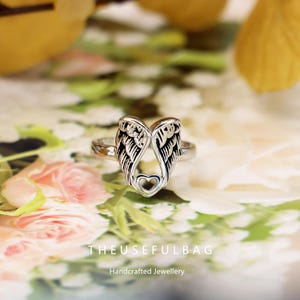 Op de afbeelding: Zilverkleurige ring met een open ontwerp van engelen vleugels en een hartvormige uitsparing aan de basis. De ring staat op een bloemenachtergrond. De tekst "THEUSEFULBAG Handcrafted Jewellery" is zichtbaar.
