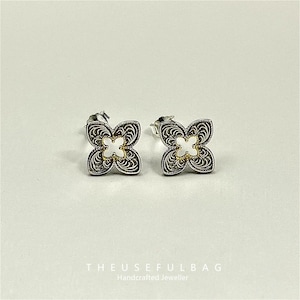 Puede incluir: Un par de pendientes de plata. Cada pendiente presenta un diseño de flor de cuatro pétalos con un patrón de filigrana. El centro de cada flor es un diseño blanco en forma de cruz con un borde dorado. El texto "THEUSEFULBAG Handcrafted Jeweller" está en la parte inferior.