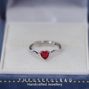May include: A silver ring featuring a heart-shaped, deep red gemstone. The band has a twisted design with small, clear stones. The ring is presented in a blue jewellery box. Text at the bottom reads: THE.USEFULBAG Handcrafted Jewellery.