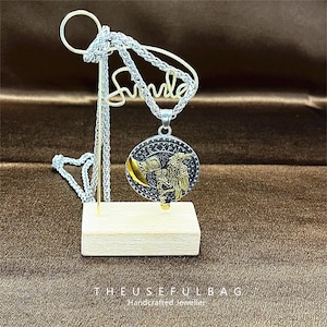May include: A silver-toned necklace with a round pendant featuring a detailed warrior design with a gold-colored axe. The pendant is displayed on a wooden block with a wire stand that has the word "smile" in cursive. The text "THEUSEFULBAG Handcrafted Jeweller" is at the bottom.