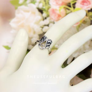 Op de afbeelding: Een zilverkleurige ring met een engelenvleugelontwerp en een hartvormige uitsparing. De ring wordt aan een vinger gedragen. De tekst "THEUSEFULBAG" en "Handcrafted Jewellery" zijn zichtbaar op de achtergrond.