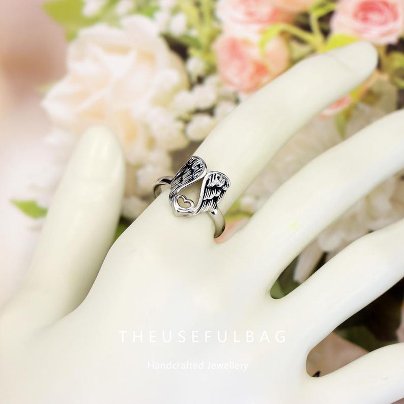 Op de afbeelding: Een zilverkleurige ring met een engelenvleugelontwerp en een hartvormige uitsparing. De ring wordt aan een vinger gedragen. De tekst "THEUSEFULBAG" en "Handcrafted Jewellery" zijn zichtbaar op de achtergrond.