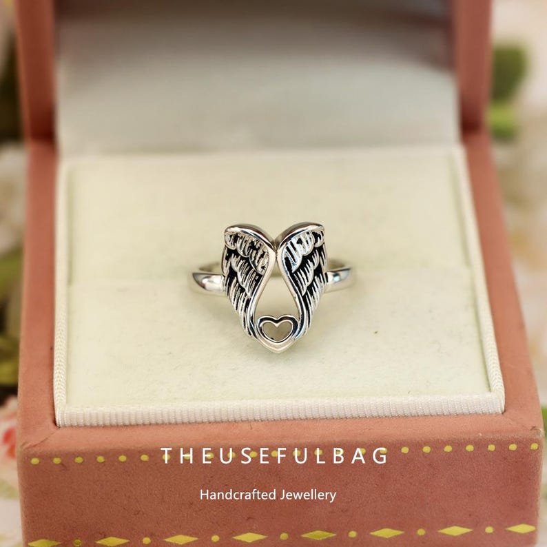 Op de afbeelding: Een zilveren ring met een engelenvleugelontwerp en een hartvormige uitsparing. De ring wordt gepresenteerd in een roze sieradendoosje. De tekst "THEUSEFULBAG" en "Handcrafted Jewellery" zijn zichtbaar op de doos.