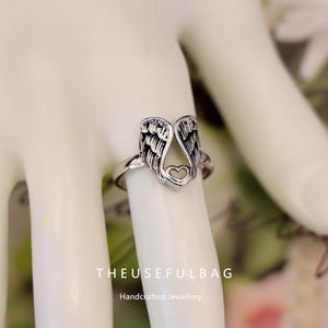 Op de afbeelding: Zilverkleurige ring met een open vleugelontwerp en een hartvormige uitsparing. De ring wordt aan een vinger gedragen, waarbij de gedetailleerde vleugels worden benadrukt. De tekst "THEUSEFULBAG" en "Handcrafted Jewellery" zijn zichtbaar.