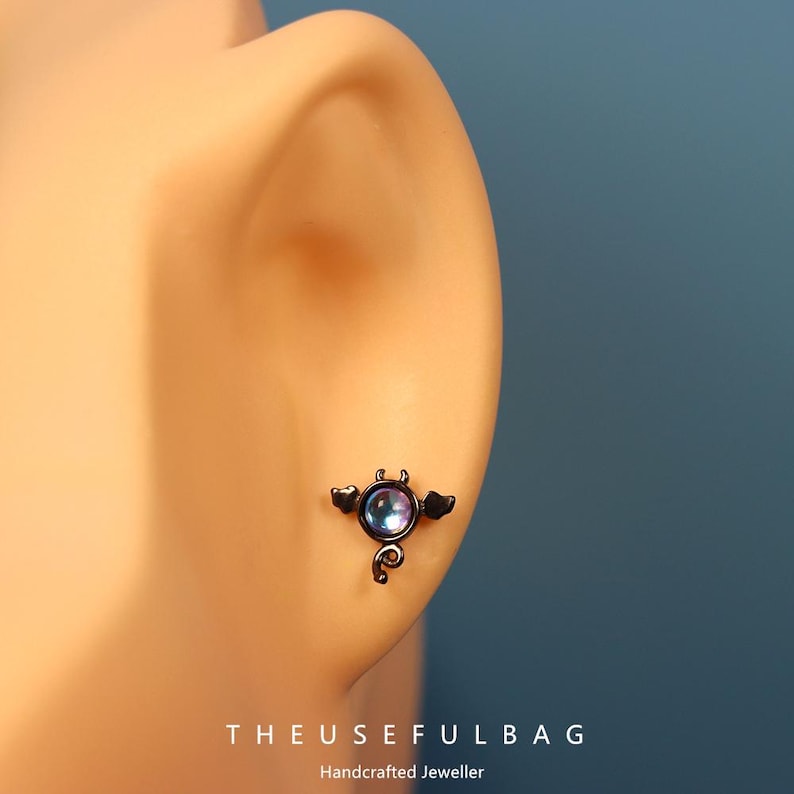 May include: A black stud earring with a devil design, featuring a round, iridescent stone at the center. The earring has small wings, horns, and a curled tail. The text "THEUSEFULBAG" and "Handcrafted Jeweller" are at the bottom.