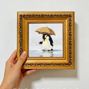 May include: A square oil painting in an ornate gold frame depicting a penguin standing on a light blue surface while holding a brown umbrella. The painting features a white background and the text THEUSEFULBAG is printed at the bottom of the image.